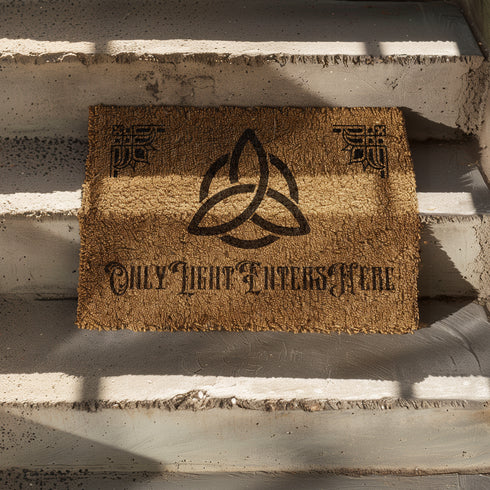 Coir doormat featuring large triple moon and triquetra symbol in center, surrounded by celestial designs including stars and moon phases, all in dark brown on natural tan background.